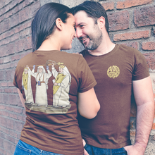 Load image into Gallery viewer, A couple wearing matching brown graphic t-shirts posing against a red brick wall. The woman shows the back of her shirt with a detailed illustration, while the man shows a gold circular logo on the chest.