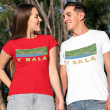Load image into Gallery viewer, A man and a woman holding hands, standing outdoors in front of trees and a fence. The woman wears a red t-shirt with a landscape graphic and the text 'Y BALA', while the man wears a white t-shirt with the same graphic.